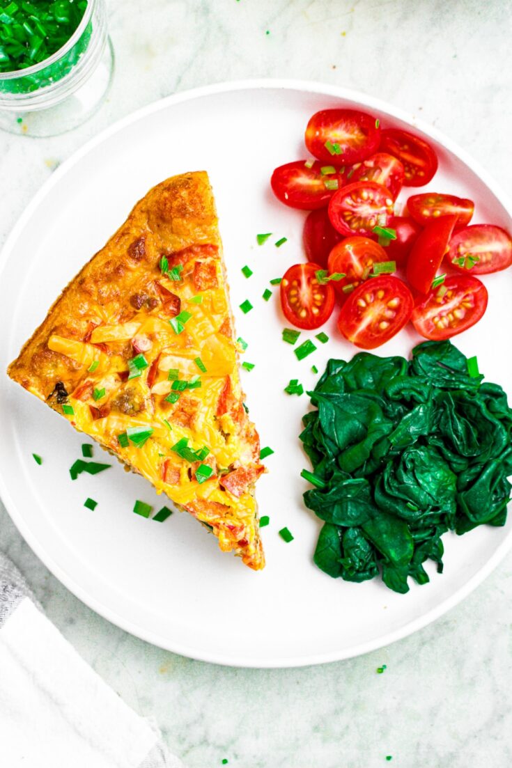 Overhead photo of a round white plate with a slice of vegan egg casserole, spinach, and sliced grape tomatoes sprinkled with minced green onion