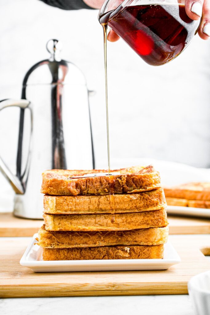 Head on photo of a stack of vegan french toast with a hand pouring maple syrup on top
