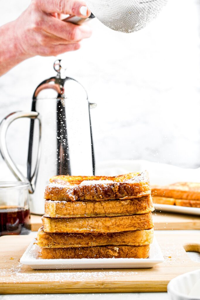 Head on photo of a stack of eggless French toast with a hand sifting powdered sugar on top