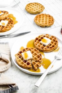 Head on photo of two plates with waffles, vegan butter, and syrup on them. More waffles are stacked in the back right corner.