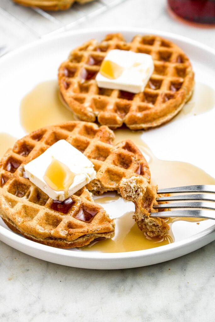 Head on photo of a plate with two waffles topped with vegan butter and syrup. A fork is pulling a piece of the front waffle away.