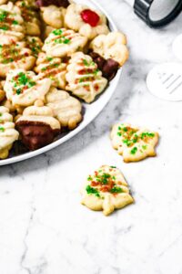 Overhead photo of 2 tree shaped spritz cookies with green and red sprinkles sit in front of a large plate of more spritz cookies