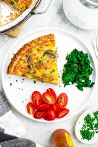 Overhead photo of a slice of vegan quiche on a round white plate with spinach, tomato slices, and minced green herbs