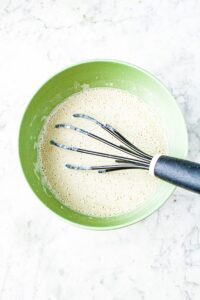 Overhead photo of a small mixing bowl with foamy white liquid in it and a whisk going in