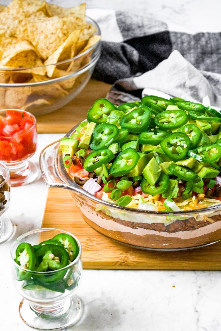 Head on photo of a glass pyrex container with layered vegan taco dip topped with sliced jalapenos. There is a bowl of tortilla chips behind the dip.