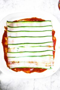 Overhead photo of zucchini strips layered on top of sauce in a square casserole dish