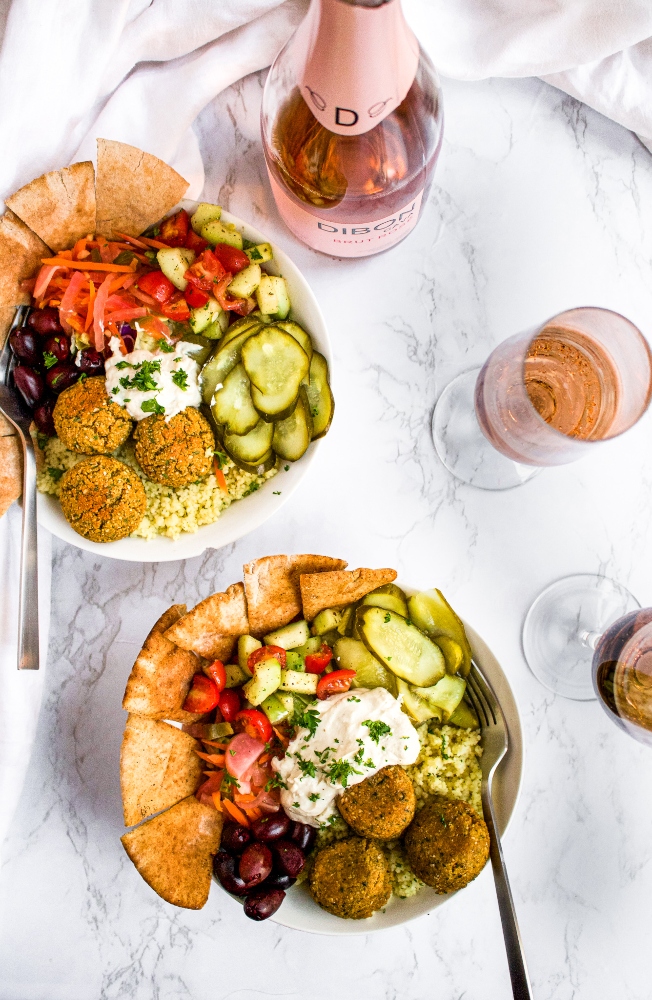 Overhead photo of two falafel bowls with pickles, pita chips, hummus, cucumber tomato salad and herbs