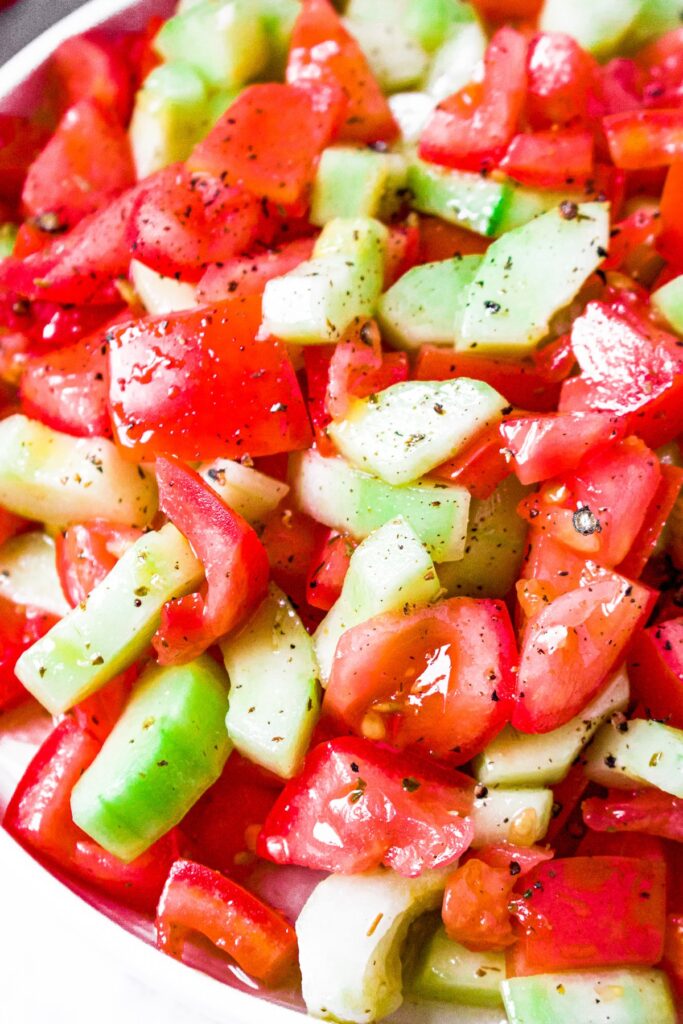 A very close up photo of cucumber salad with tomatoes, Italian dressing, and topped with freshly ground black pepper