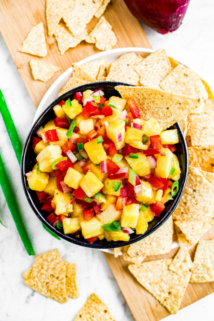 Overhead photo of a bowl of spicy and sweet pineapple salsa with a chip standing in it. The bowl is surrounded by more tortilla chips and a couple scallions.