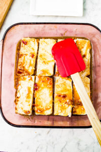 A spatula spreads teriyaki mayo on vegetarian chicken pieces in a square clear baking dish