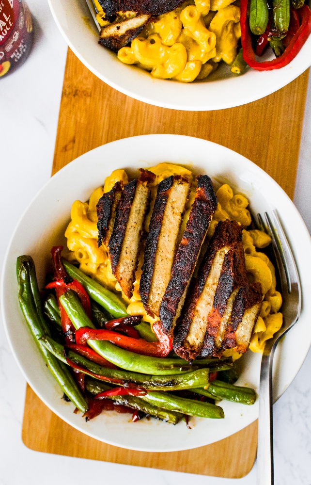 Overhead photo of a bowl with a sliced barbecue tofu cutlet on top of vegan mac and cheese and next to green beans and peppers