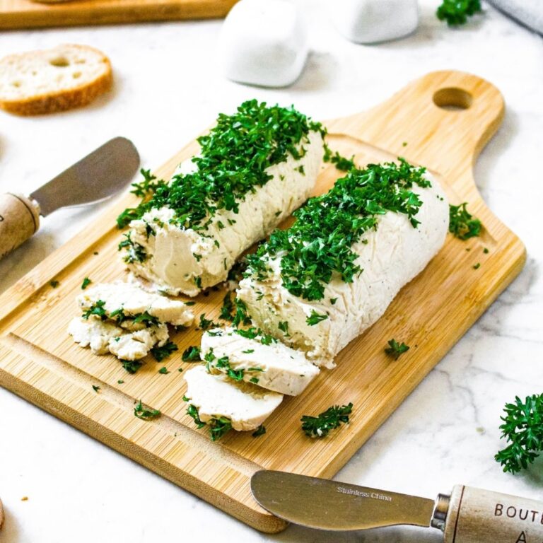 Head-on view of two tubes of vegan goat cheese on a wooden cutting board topped with fresh minced parsley. There are slices of baguette surrounding the board along with cheese knives, and salt and pepper shakers