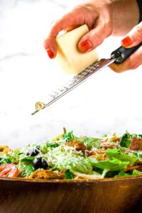 Head-on photo of a big green salad with a hand grating vegan parmesan on top