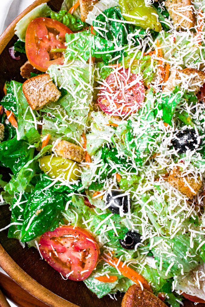 Overhead zoomed in photo of a big green Italian salad with pepperoncini, black olives, tomatoes, croutons, and dairy-free parmesan