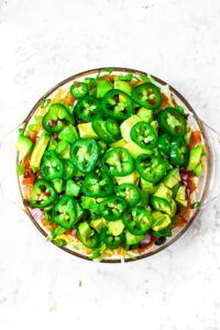 Overhead photo of a round clear glass dish with vegan nacho dip topped with diced avocado and sliced jalapenos