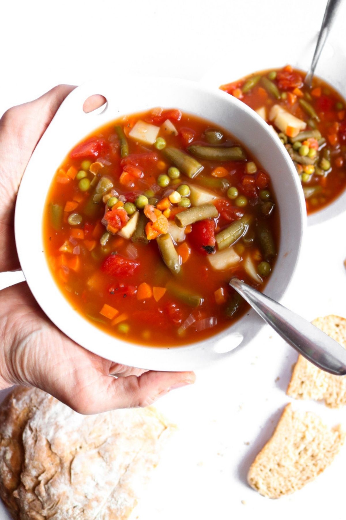 Overhead view of vegetarian vegetable soup in a round white bowl with a spoon digging into it and bread slices off to the side.