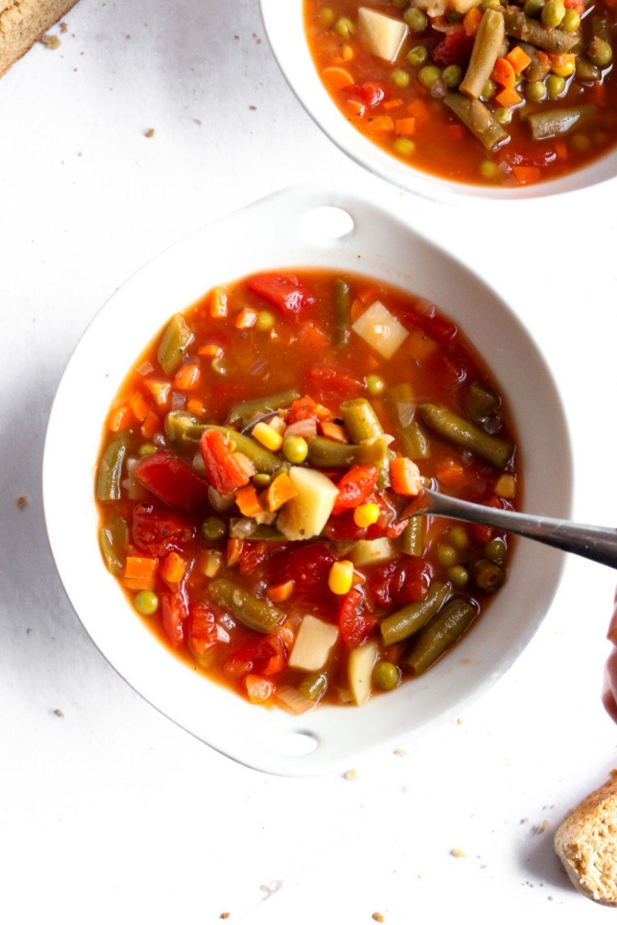 Overhead view of vegetarian vegetable soup in a round white bowl with a spoon digging into it and bread slices off to the side.