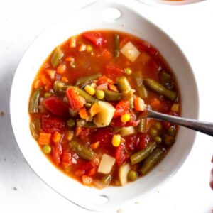 Overhead view of vegetarian vegetable soup in a round white bowl with a spoon digging into it and bread slices off to the side.