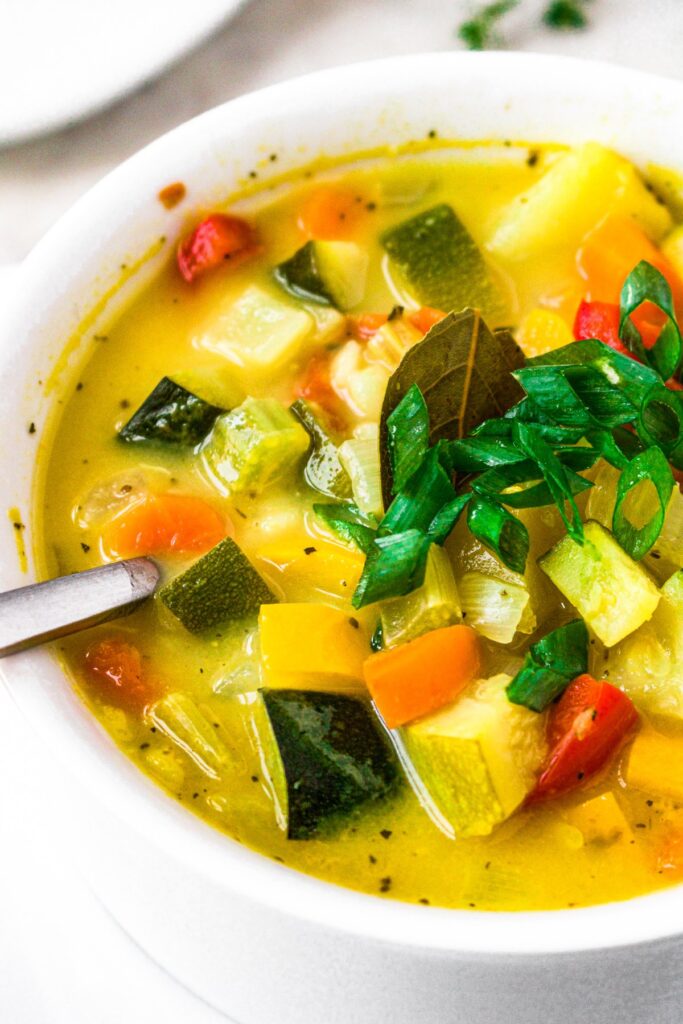 Close up photo of a bowl of squash and zucchini vegetbale soup with a vegan chicken broth topped with scallions in a white bowl with a spoon digging into it