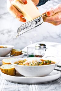 Head on photo of hands grating vegan parmesan over a bowl of minestrone soup
