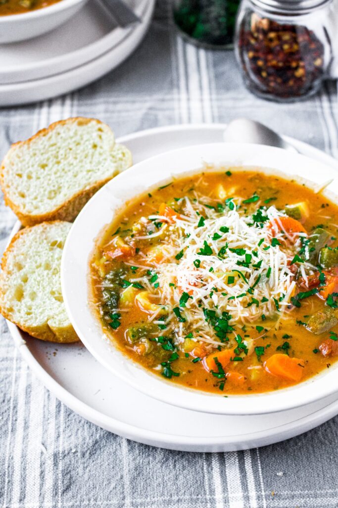 Overhead photo of a round white bowl of minestrone vegetable soup on a plate next to baguette slices