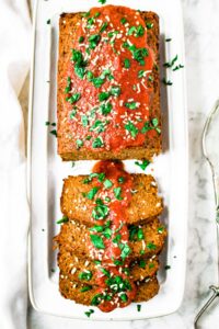 Overhead photo of a half-sliced vegetarian meatloaf on a rectangular white plate topped with tomato sauce, minced herbs, and dehydrated onions.