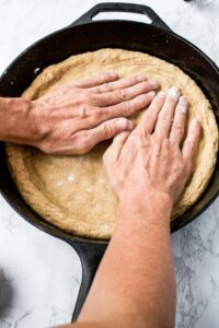 Overhead photo of two hands pressing a wheat pizza crust into a cast iron pan