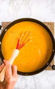 Overhead photo of a hand with a whisk stirring a large pot of pumpkin soup with coconut milk