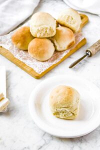 Overhead photo of a dinner roll on a small round white plate in front of a wooden cutting board with a pile of dinner rolls on it