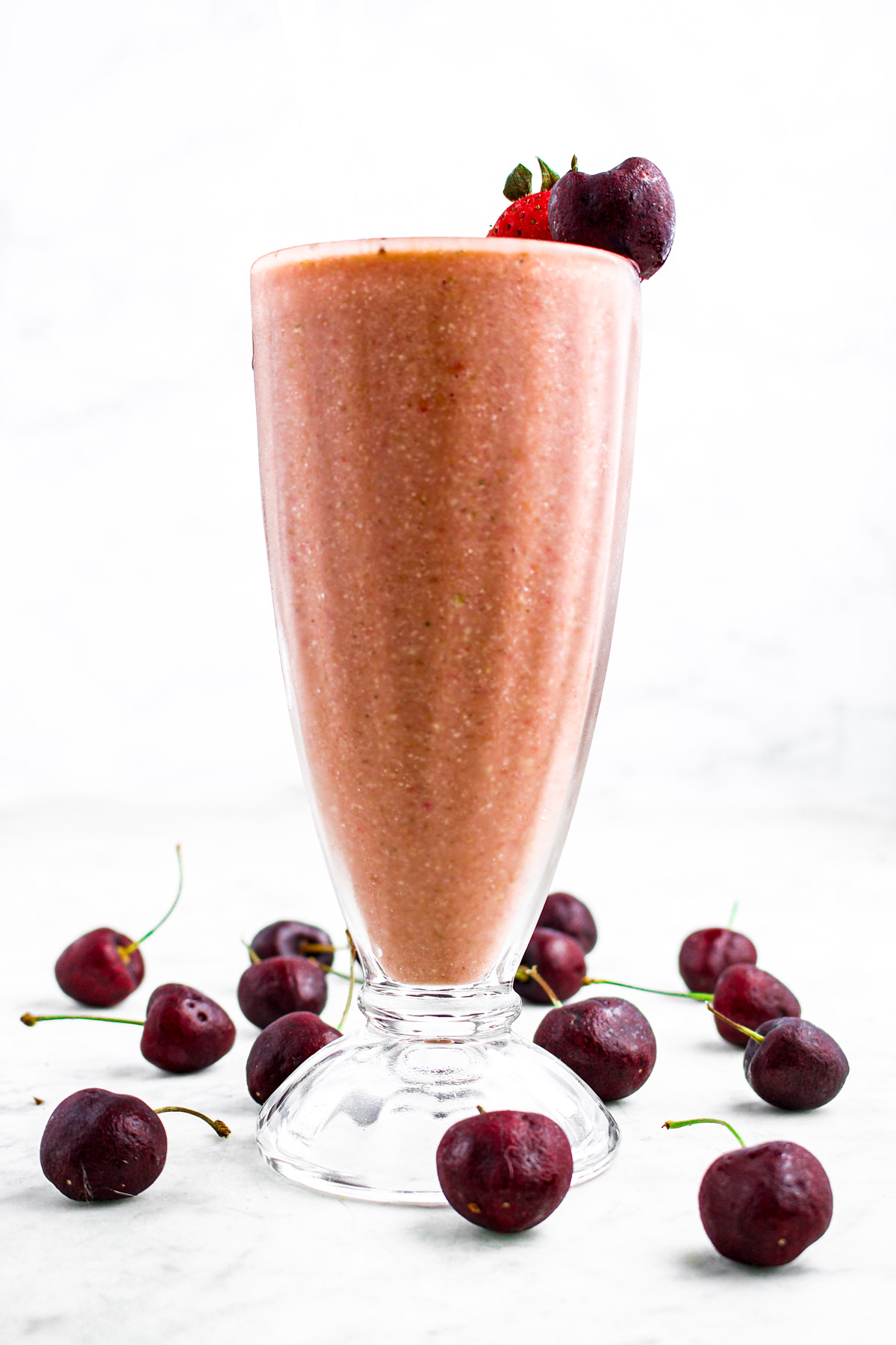 Head on photo of a glass of pink smoothie with a strawberry on the rim surrounded by fresh cherries