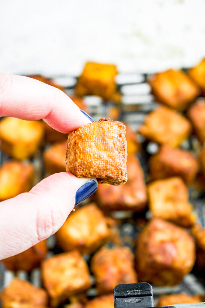 A hand with blue nails lifts a golden brown tofu cube out of an air fryer basket