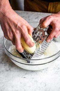 Overhead photo of two hands grating a peeled potato into a glass bowl of water