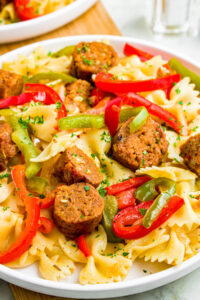 Close up photo of a pile of farfalle pasta topped with sliced seitan sausages, onions, and peppers