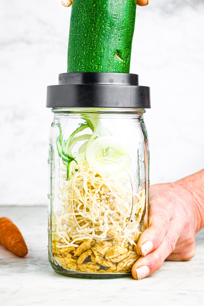 Head on photo showing a zucchini being spiralized into a large clear mason jar