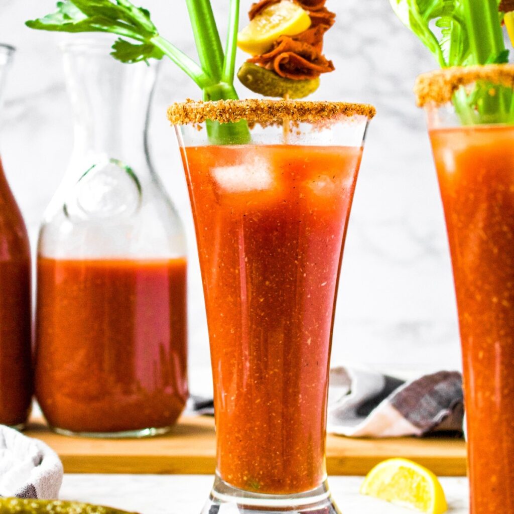 Head on photo of a tall glass of vegan Bloody mary garnished with a celery stalk and skewer with vegan meats, lemon, and olives