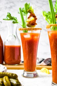 Head on photo of a tall bloody mary glass with a spicy salted rim in front of a pitcher of bloody marys