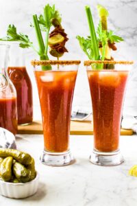 Head on photo of two tall glasses of vegetarian bloody marys with a celery stick sticking into them and skewers with olives and vegan meats