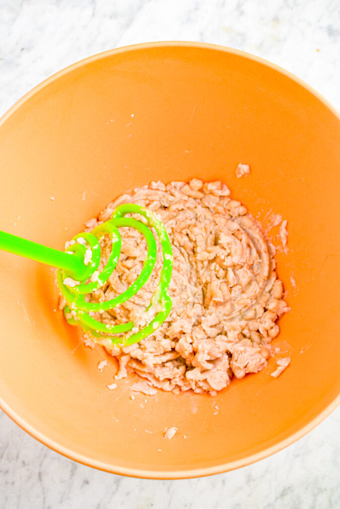 Overhead view of partially mashed white beans in a large orange mixing bowl with a potato masher