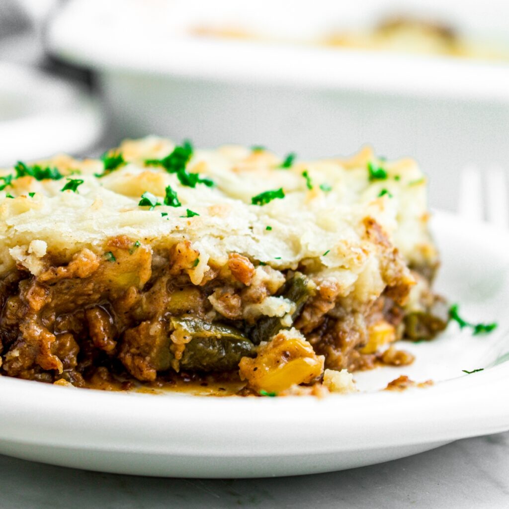 Head on close up view of a slice of vegan shepherd's pie on a round white plate topped with green herbs