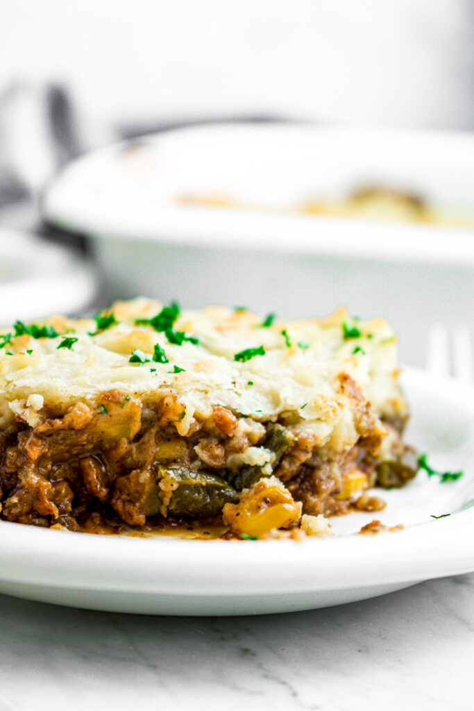 Head on close up view of a slice of vegan shepherd's pie on a round white plate topped with green herbs