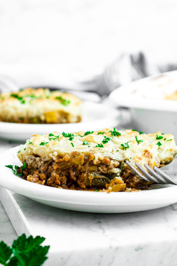 Head on close up view of a square of dairy free shepherds pie on a round white plate topped with green herbs next to a fork