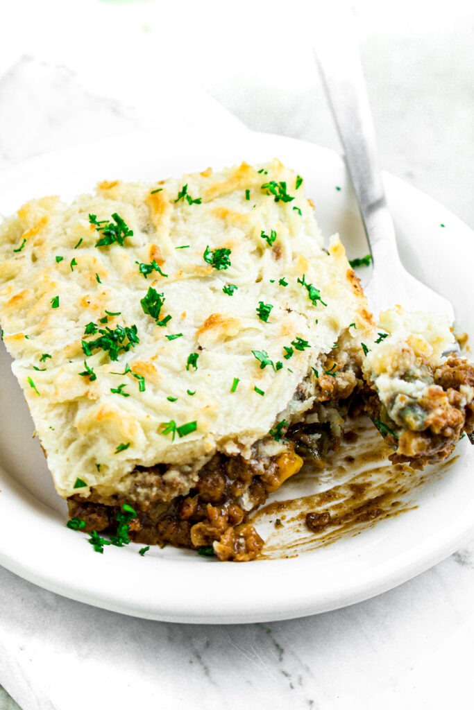 Overhead close up view of a slice of vegan cottage pie on a round white plate topped with green herbs
