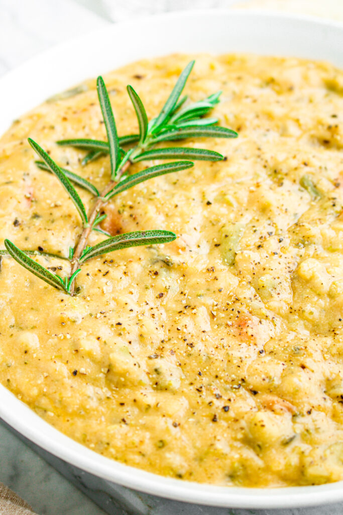 Overhead zoomed in view of a round white bowl filled with creamy old fashioned vegetable soup with a sprig of rosemary sticking out of it on the left side