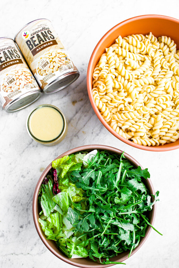 Overhead view of a large bowl of pasta, another bowl of arugula and greens, a small bowl of creamy lemon dressing, and two cans of white beans