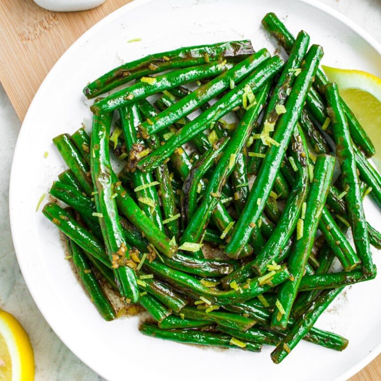 Overhead close up view of a round white plate piled high with lemon pepper green beans and a lemon wedge to the right side
