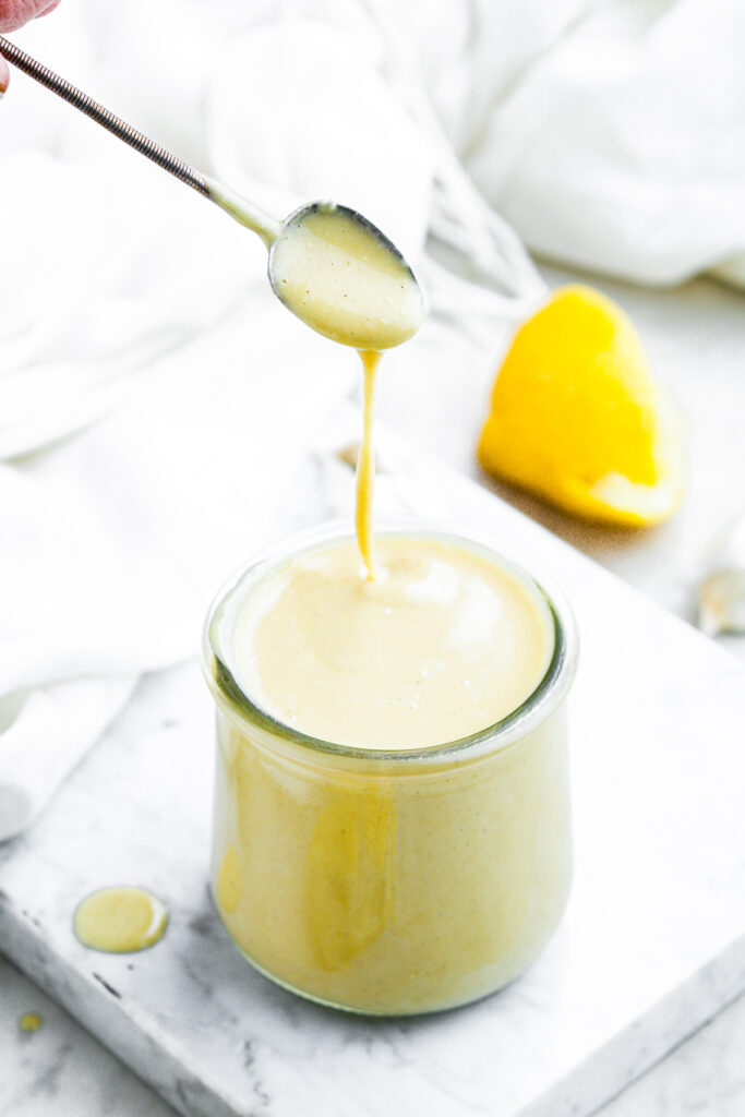 Head on view of a spoon drizzling lemony vinaigrette into a clear glass jar with a sliced lemon behind it on a marble countertop