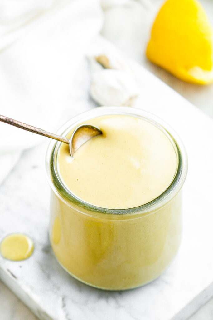 Overhead view of a spoon diving into a glass jar filled with creamy vegan lemon salad dressing with sliced lemons behind it