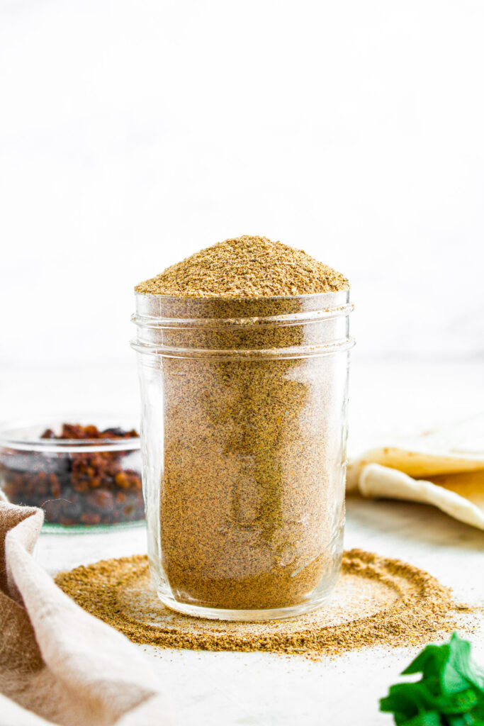 Head on view of a clear glass jar of DIY taco seasoning with some spilling out creating a circle around the jar