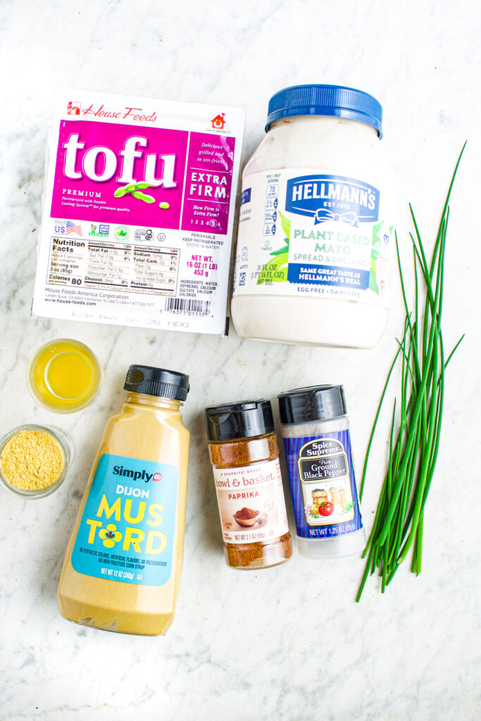 Overhead view of ingredients on a marble countertop including vegan mayo, tofu, dijon mustard, pickle juice, tofu scramble seasoning, pepper, paprika, and chives