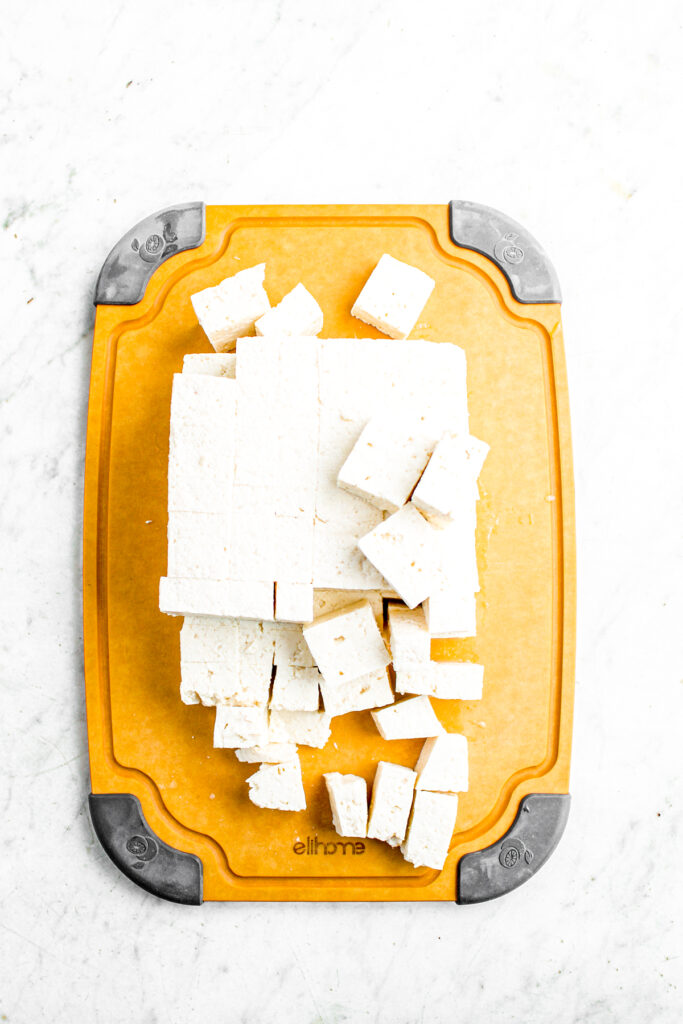 Overhead view of a diced block of tofu with half of the block scattered about a wooden cutting board